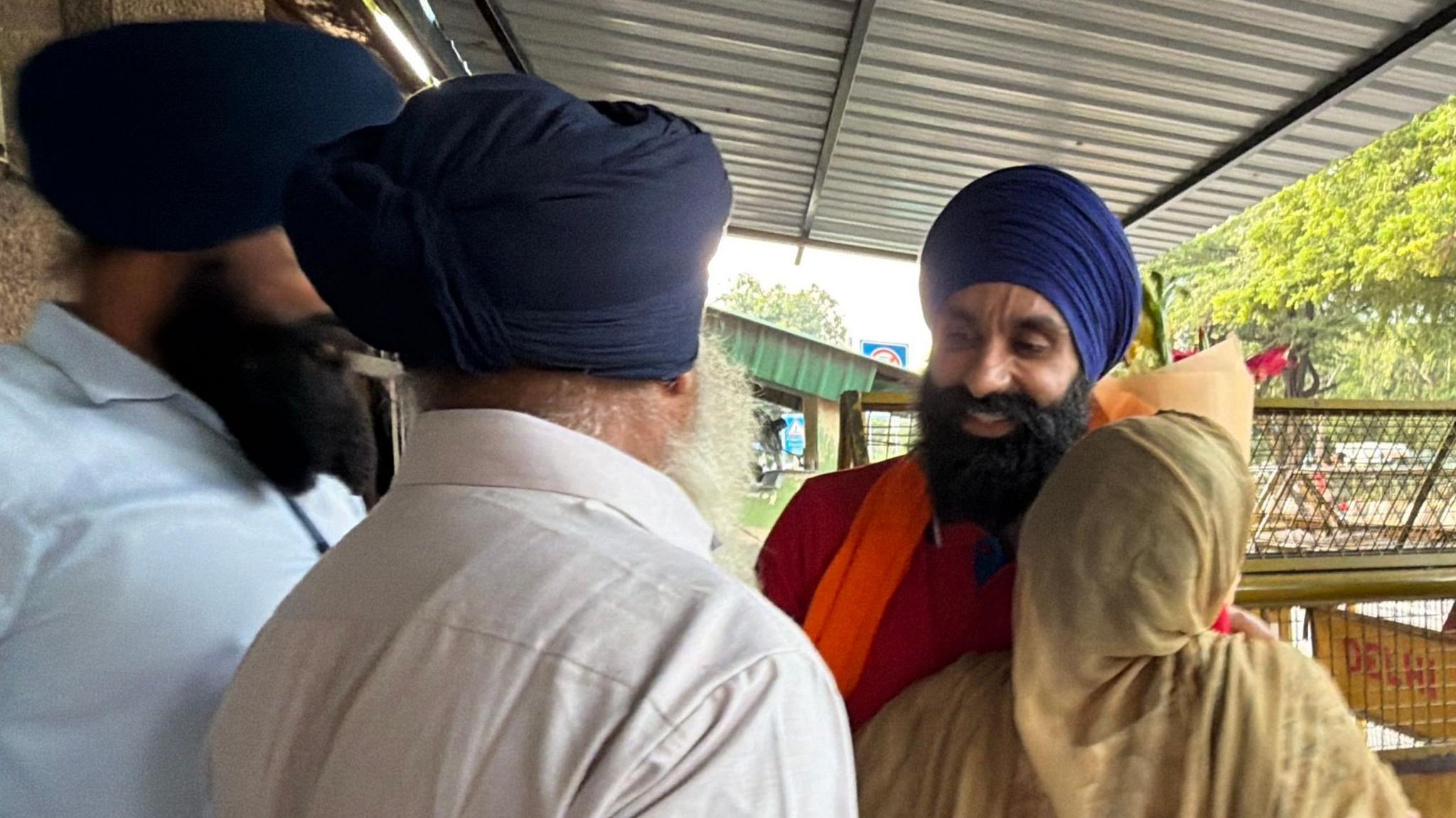 Jagtar Singh Johal, wearing a red top with an orange garment around his neck, blue turban and dark beard, being embraced by family members outside the jail in Delhi.