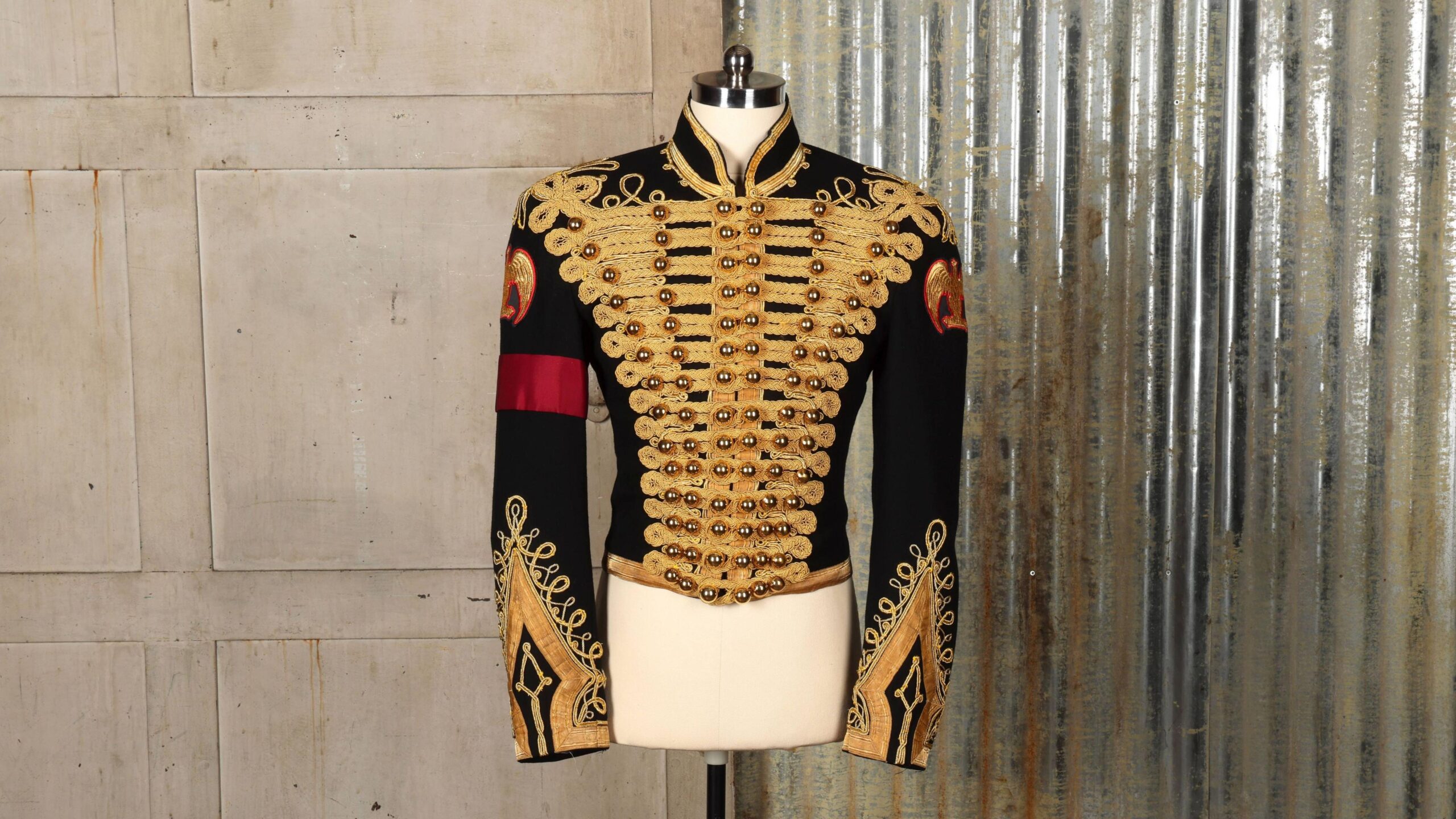 A black and gold military-style jacket, on a white mannequin, in front of a wall with a curtain to the right. It has a red arm band on the right arm.