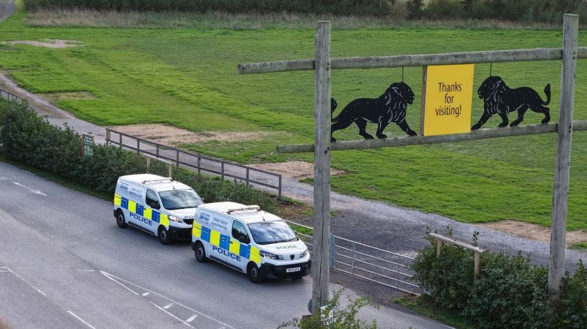 Two police vans on a road at the entrance to the park. There is a suspended yellow sign which says 'thank you for visiting' and two black lion signs at either side.