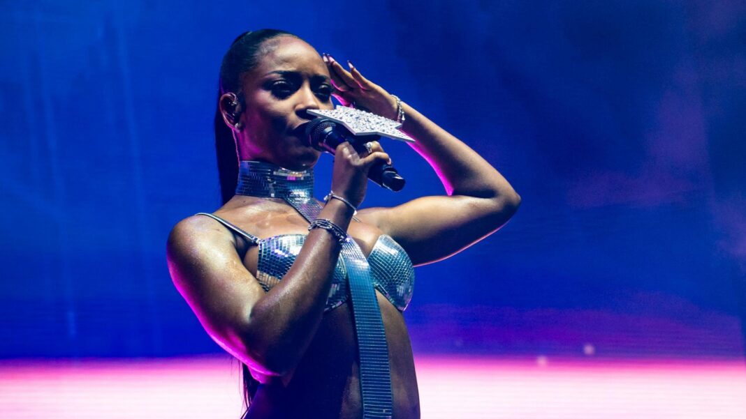 Afrobeats singer Ayra Starr on making history at her NFL half-time show Ayra Starr performs during All Points East in London. She wears a sparkly bra top and holds a microphone with a large star on it.