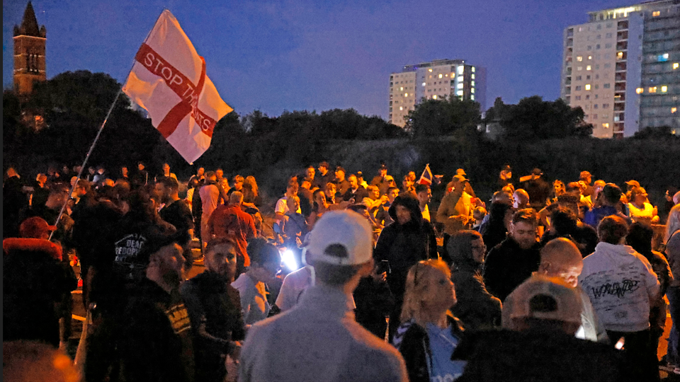 Demonstrators are standing around, it's quite dark and there are a couple of tall buildings in the background. A red and white flag reads Stop the Boats