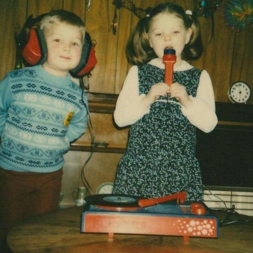 Vintage photograph of two children indoors beside a record player, with one child wearing large ear defenders and the other holding a toy microphone in front of a wood-panelled wall.