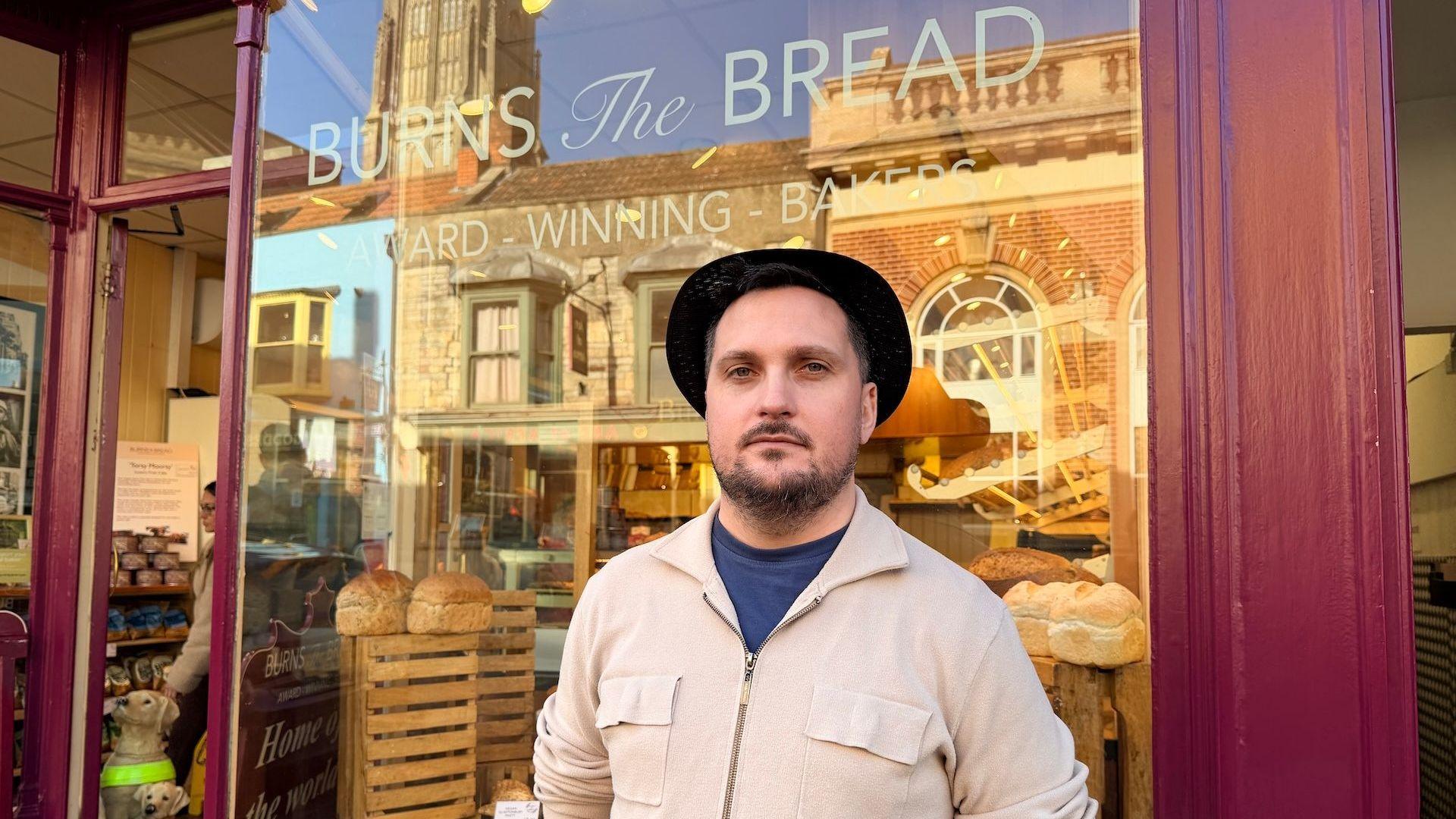 A man is stood wearing a beige zip-jacket with a blue top underneath, as well as a rimmed black-hat. Behind him is the red-framed shop window of a bakery, with bread in the window.