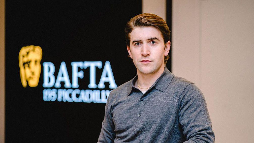 Callum Scott Howells in a grey collared jumper, pictured in front of the Bafta headquarters in Piccadilly. His dark hair is slicked back, and he looks directly at the camera.