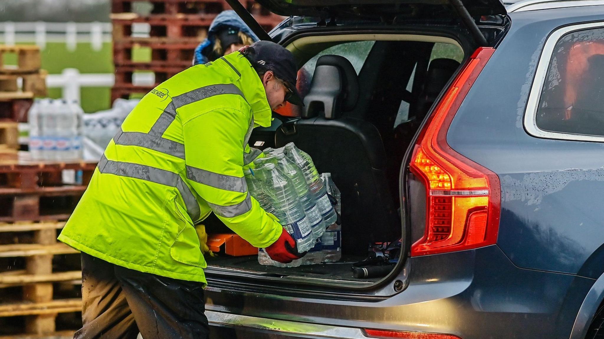 A man in a high-vis jacket, black trousers and a cap, wearing red gloves loads bottles of water into the boot of a blue SUV. In the background are pallets with more bottled water.