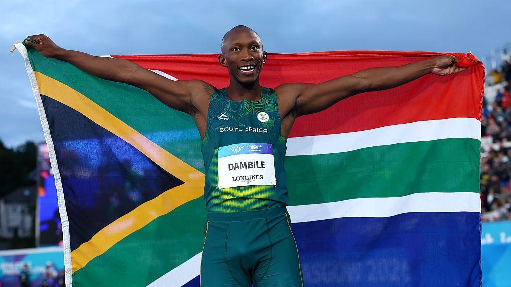 Sinesipho Dambile wearing a green South Africa sleeveless running vest and shorts, with a white name bib on his stomach, stretches his arms wide to either side of his body to raise a South African flag behind him.