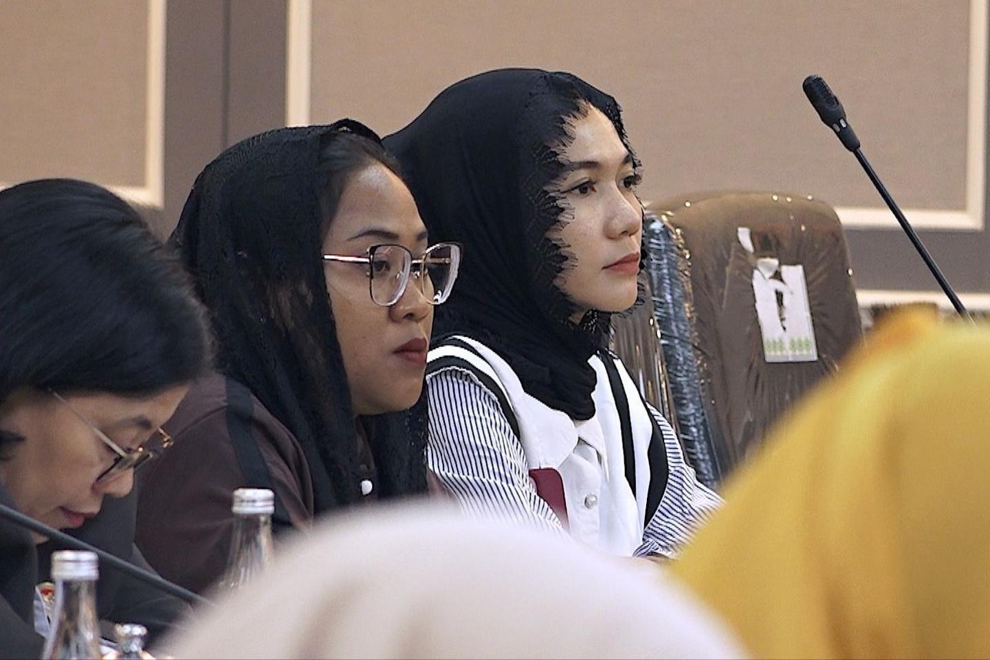 Three women seen at the parliamentary hearing into Bechi's release. Two wearing dark head coverings are in focus near the centre of the image, seated beside microphones and water bottles, while other participants are partially visible in the foreground. The room has neutral-coloured walls and conference-style seating. A microphone stand is positioned above the table.