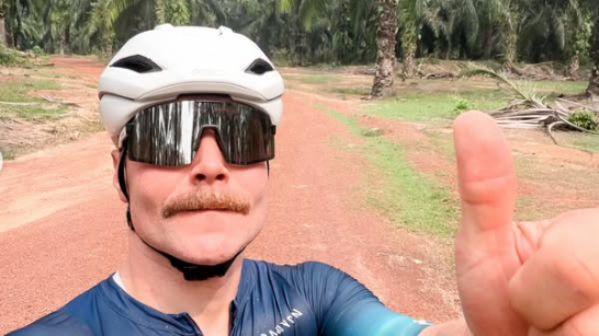 Valterri Bottas, wearing a cycling top and helmet with sunglasses, gives a thumbs-up in a selfie photo with a dirt track and palm trees in the background