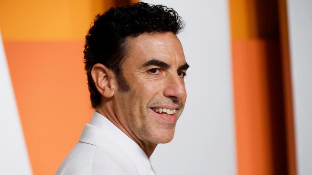 Sacha Baron Cohen smiles while wearing a white suit at the Vanity Fair Oscars party after the 97th Academy Awards, in Beverly Hills, California in March 2025.