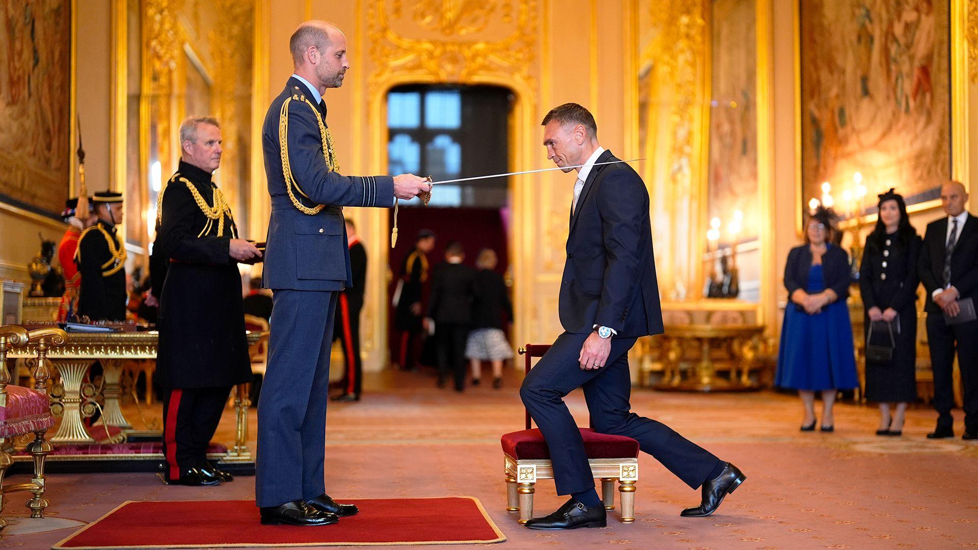 The Prince of Wales knights Sir Kevin Sinfield with a sword at an investiture in a grand, gold room at Windsor Castle.
