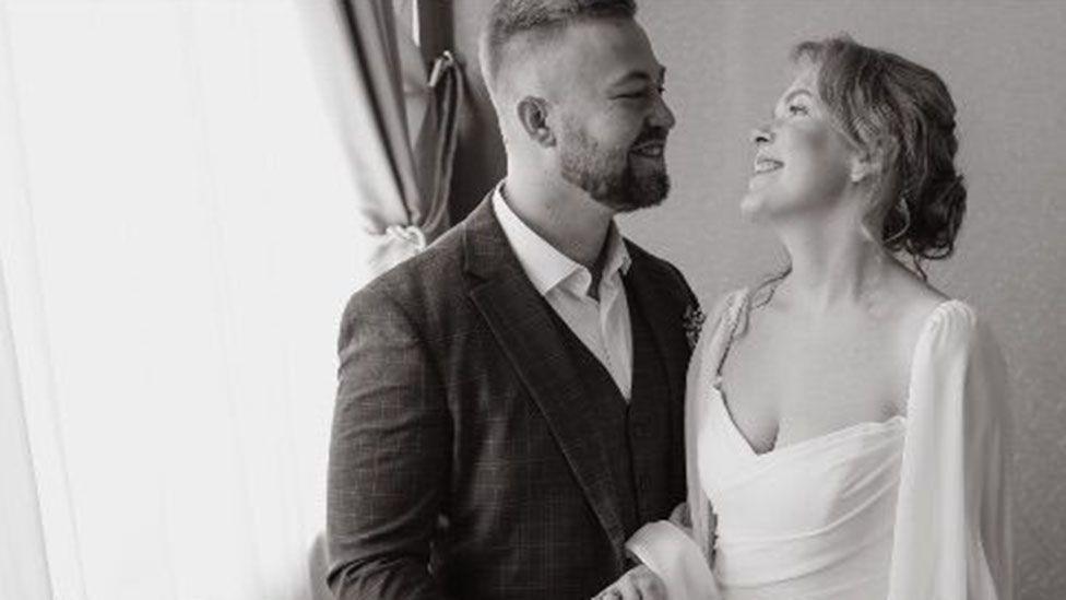 Nathan Stobbart is wearing a dark wedding suit and waistcoat with white shirt, unbuttoned and with no tie. He is smiling as he looks at Ksenia, who is wearing a white wedding dress with drapes from her shoulders. Nathan has short hair, shaved at the sides, with a trimmed beard and moustache. Ksenia's blonde hair is tied up in a lose bun with ringlets of hair falling down. All the colours are in black and white.