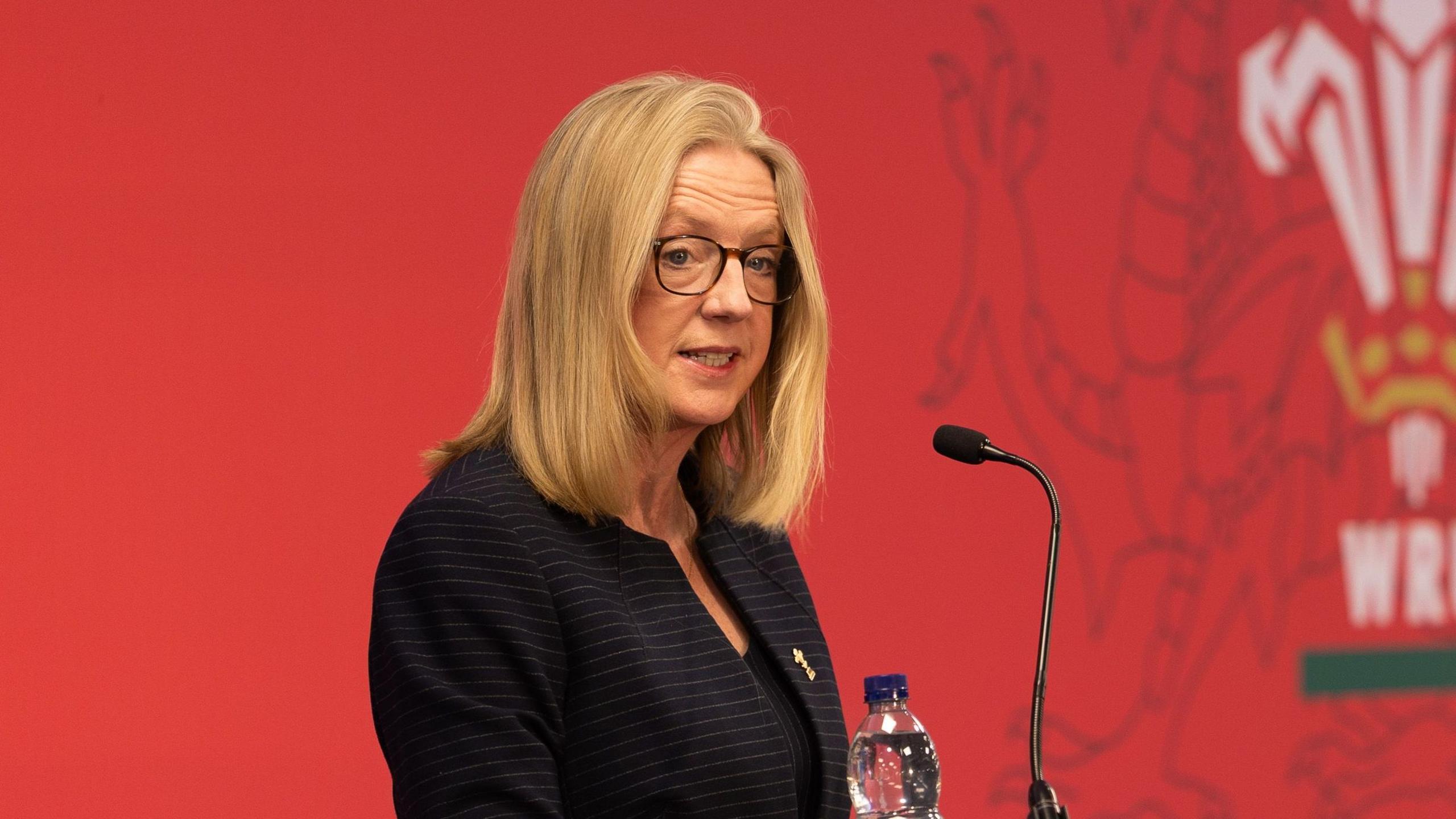 Abi Tierney wears a black jacket while standing in front of a red backdrop that includes the WRU three-feathers logo