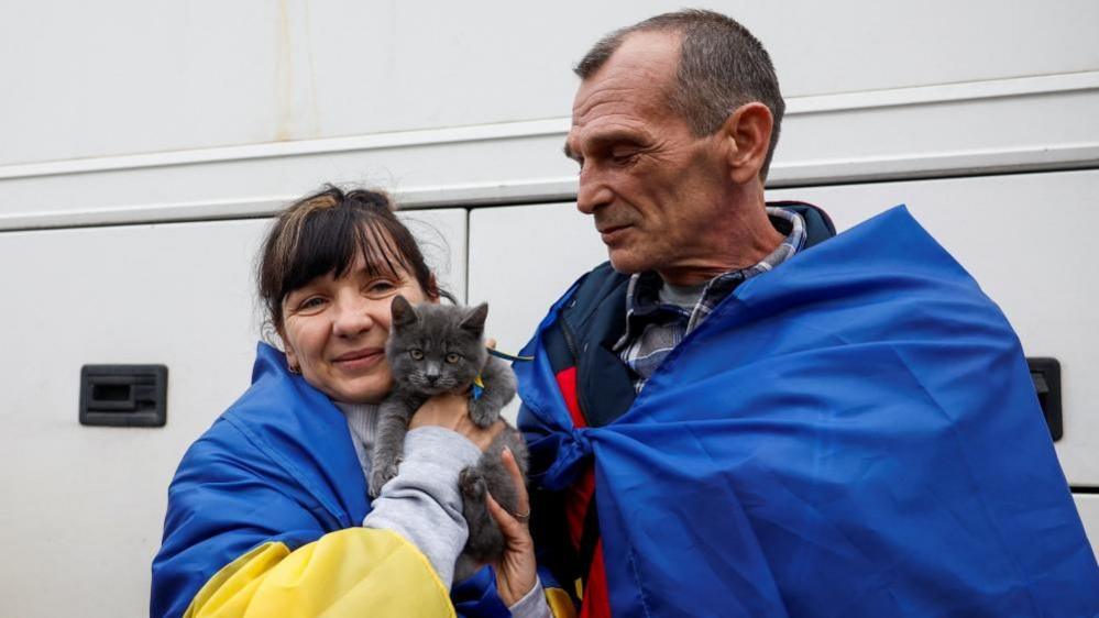 Evacuated residents pose for a photo with their cat. They are both draped in Ukraine flags