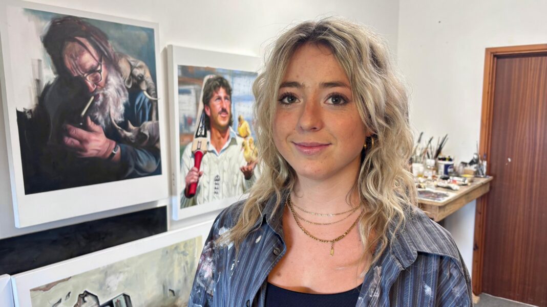 A girl with blonde wavy hair stands in a studio with paintings visible behind her. She is wearing an unbuttoned striped shirt which is covered in splashes of paint over a black top.