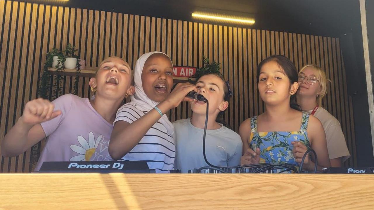 Four young girls are standing in front of DJ equipment and singing into a microphone.