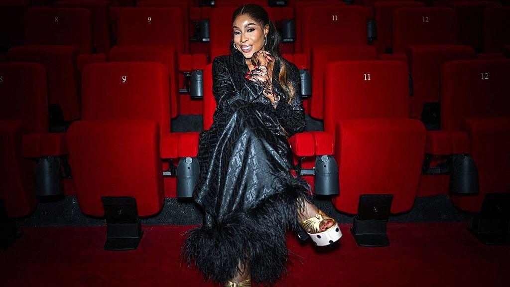 Mo Abudu seated in a red velvet cinema auditorium, wearing a patterned black long-sleeved dress with feather trim on the hem and gold platform heels.