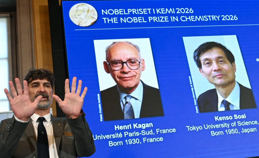 A man in a grey suit and black tie stands to the left of the picture and holds up both hands. There is a screen behind him that is titled in Swedish and English 
