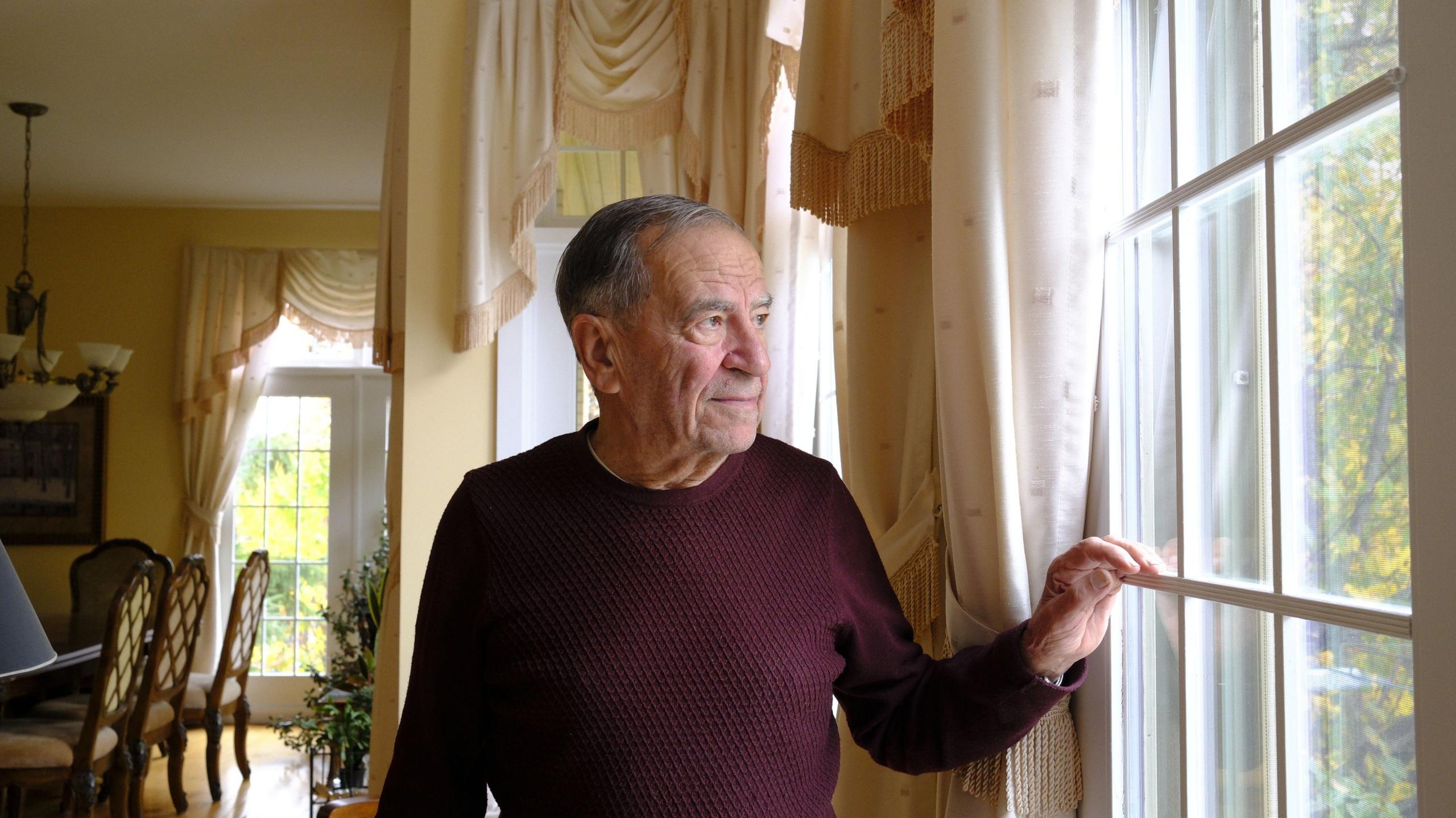 An image showing Frank Dottori inside his home, looking outside of a window. His face is illuminated by the natural light outside. He is older and has grey hair, and is wearing a deep burgundy sweater. Behind him are parts of his home dining room visible and curtains.