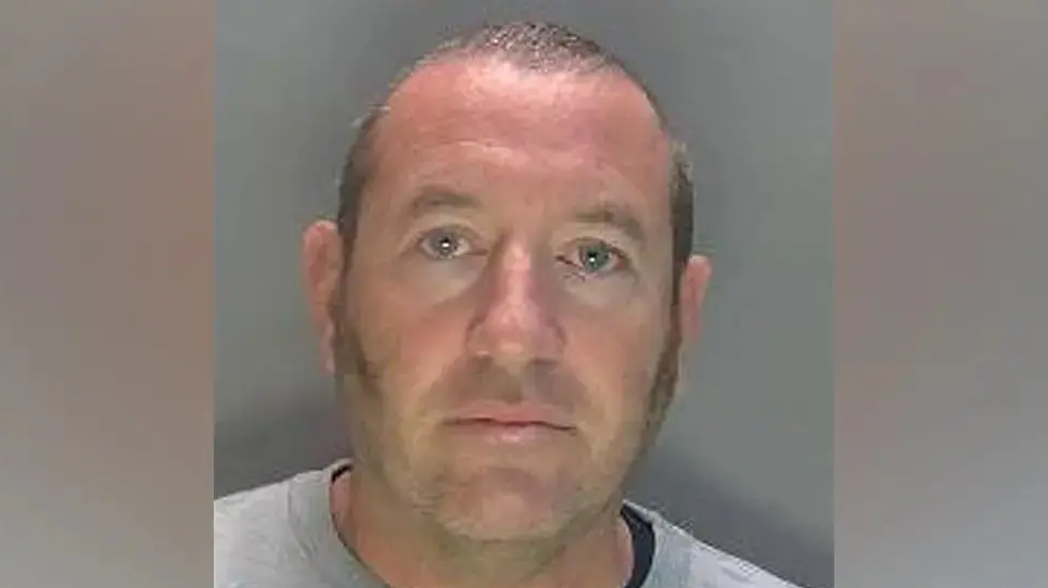 David Carrick photographed in police custody. He is wearing a grey sweatshirt and has sideburns and a near-shaven head.
