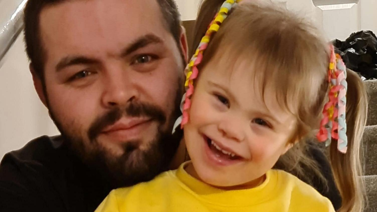 Aaron Huxter (left) with his daughter Olivia-Grace. Aaron has blue eyes, short brown hair and a bushy beard. Olivia wears a yellow jumper, has her brown hair in pigtails and smiles at the camera.