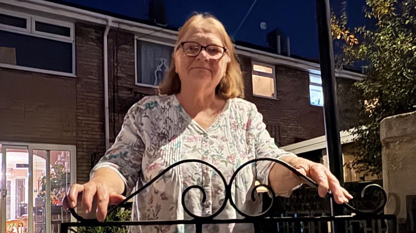A woman with light brown hair stands clutching a black wrought iron gate in front of a terraced home.