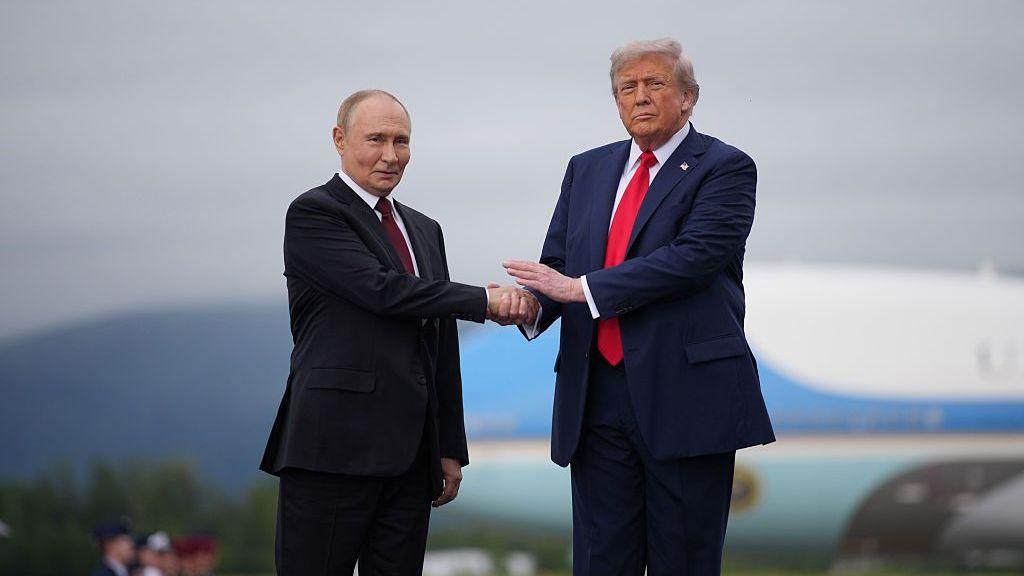 Two men shake hands as they pose for photographs. The shorter man, to the left, wears a black suit jacket and maroon tie, while the taller man, to the right, wears a dark blue suit jacket and red tie.