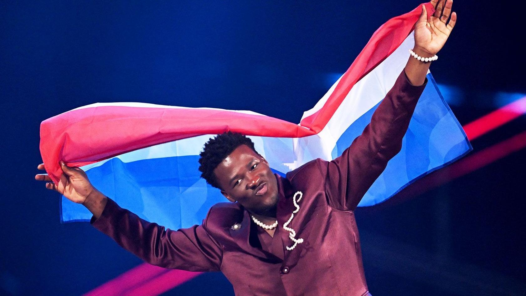 Claude Kiambe was the last Netherlands entrant in 2025 - here is wearing a burgandy suit, a pearl necklace and is holding up the Netherlands flag