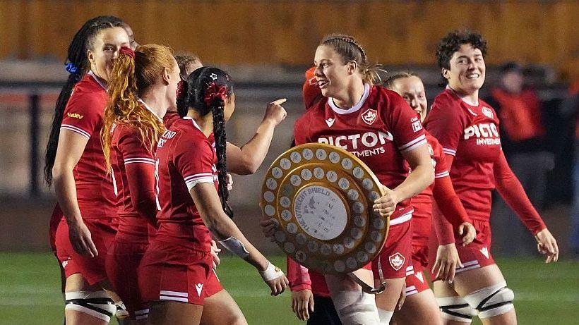 Sophie de Goede (third right) holds a brown circular trophy with white badge on it, surrounded by team-mates, all wearing red