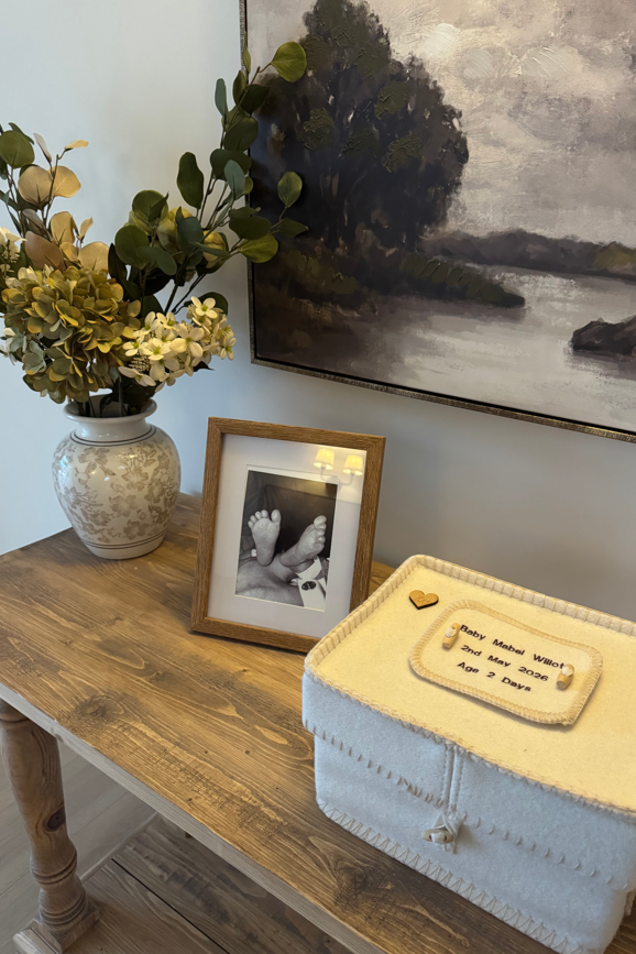 A cream felt box with Mabel's name at the top. It is sitting on a wooden console table next to a framed black and white photograph of baby Mabel's feet. Next to it is a vase of silk flowers on the wall is a large oil painting of trees around a pond or lake.