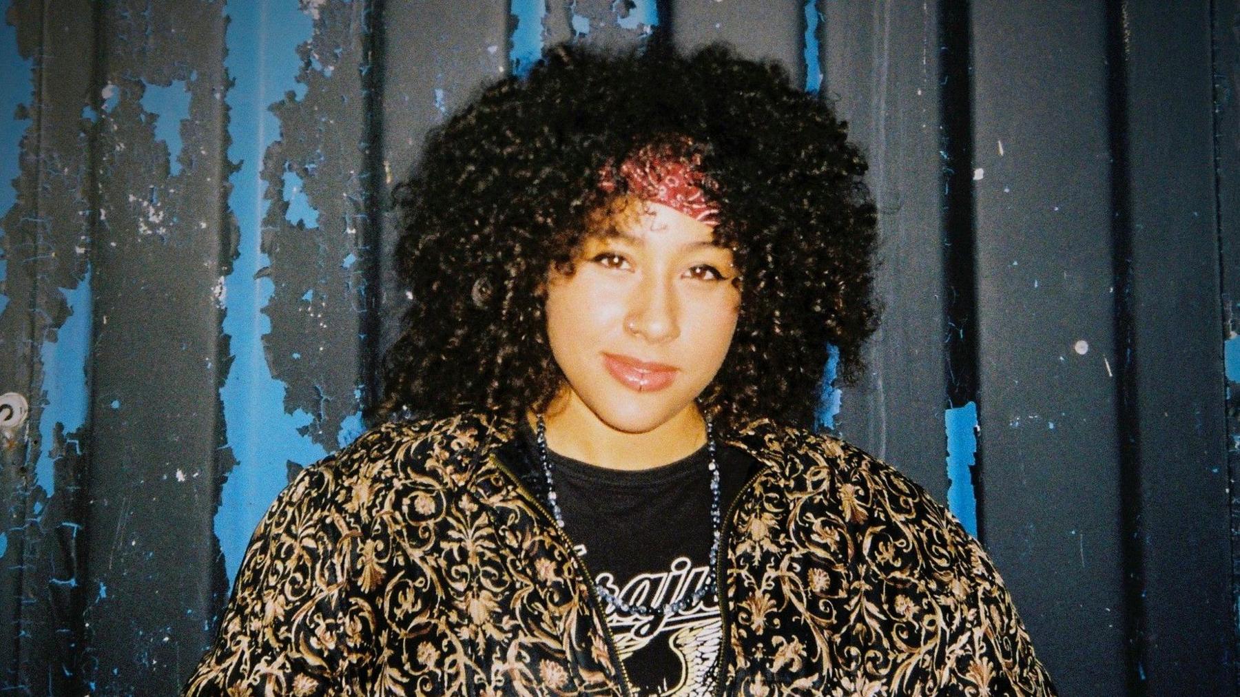 Tazmin Samuels looks at the camera as she stands in front of a metal shipping container. She has thick dark-brown curly hair and wears a black t-shirt underneath a black and gold jacket.