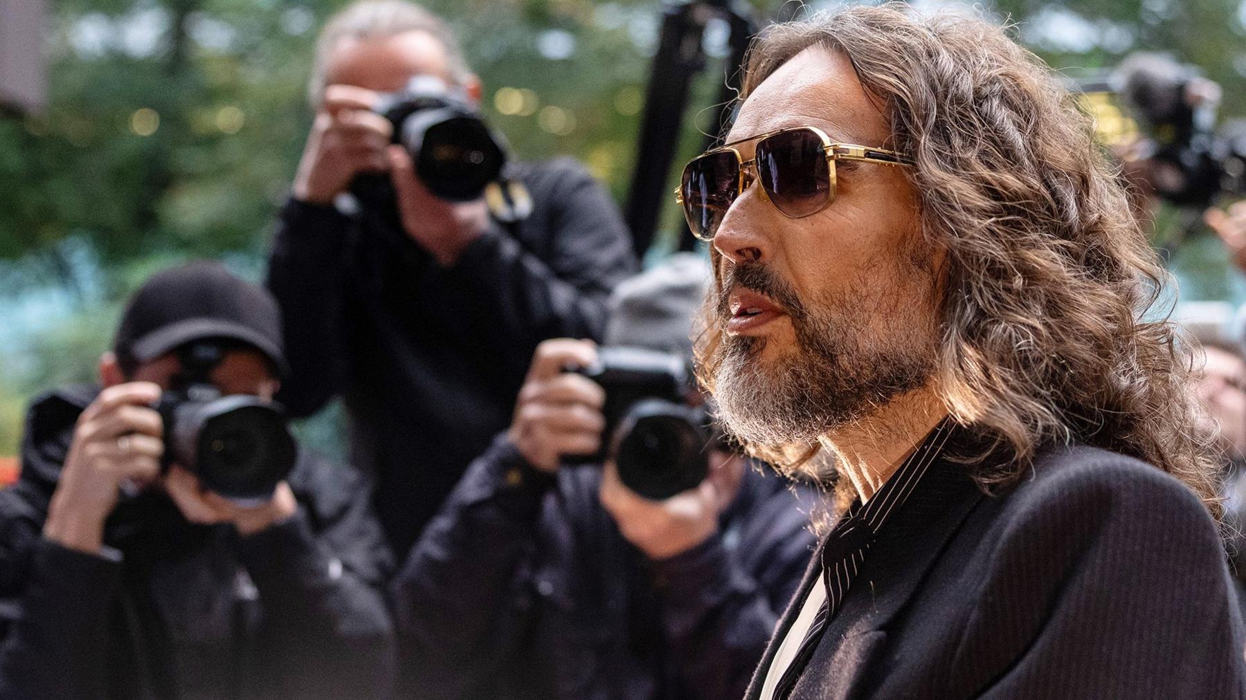 Russell Brand walking into court weraing sunglasses with three paparazzi photographers talking photos behind him