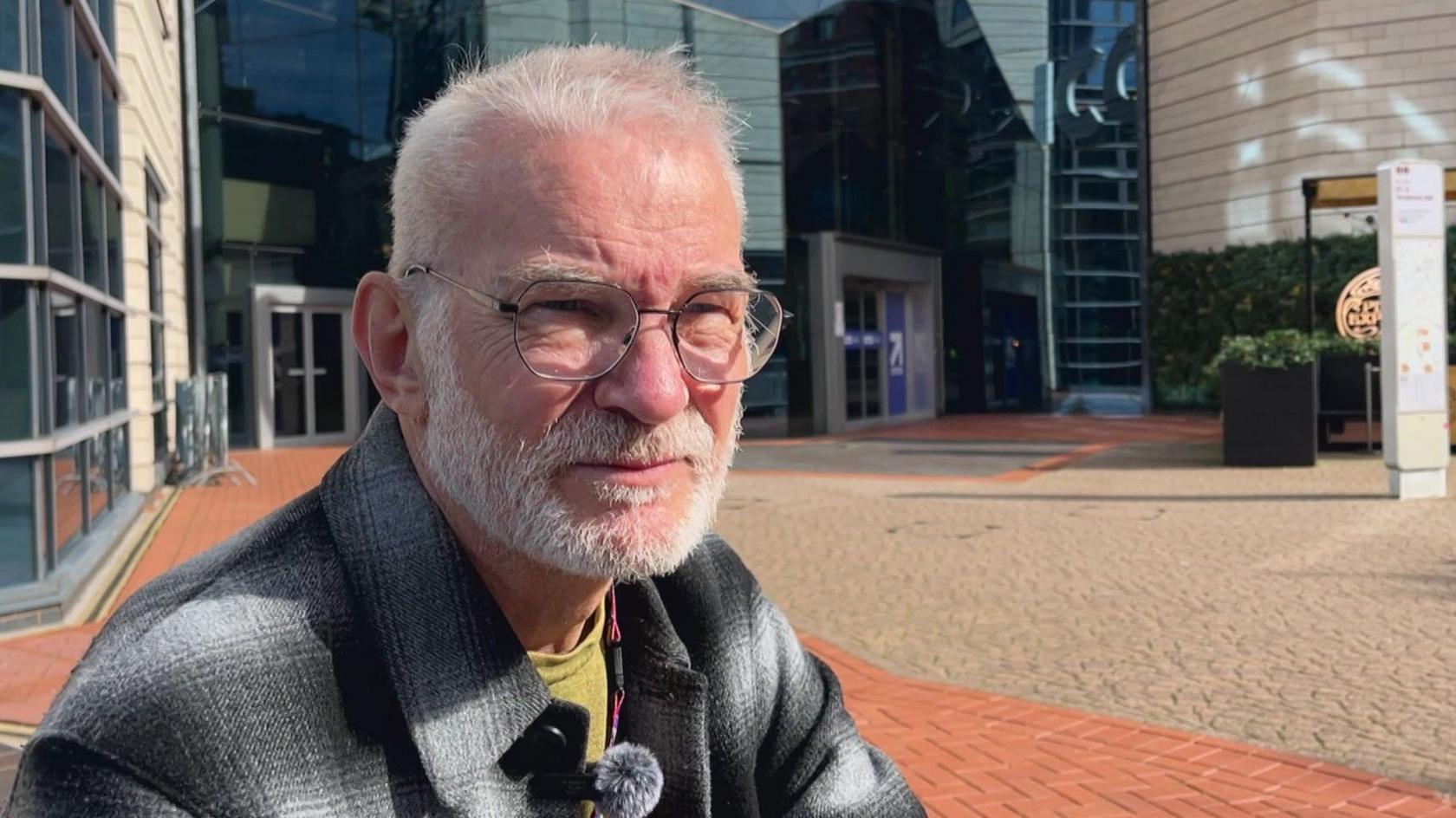 A man with grey hair and beard sits outside a modern building being interviewed. He is wearing a grey flannel shirt and mustard coloured t-shirt.