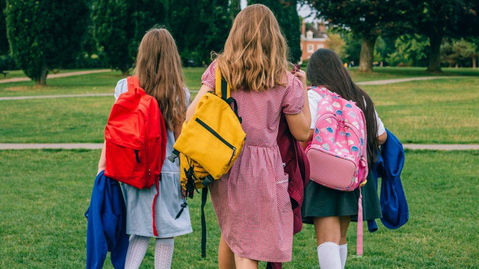 Three girls carrying school bags walk in a park with their backs to the camera