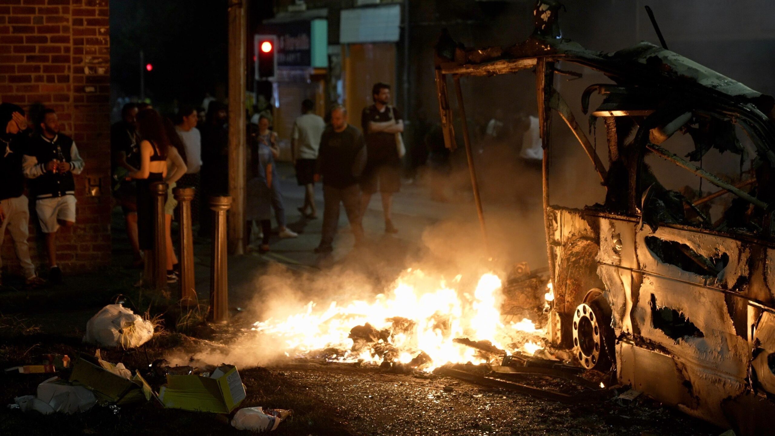 A growd gathers as a bus burns in the middle of the street.
