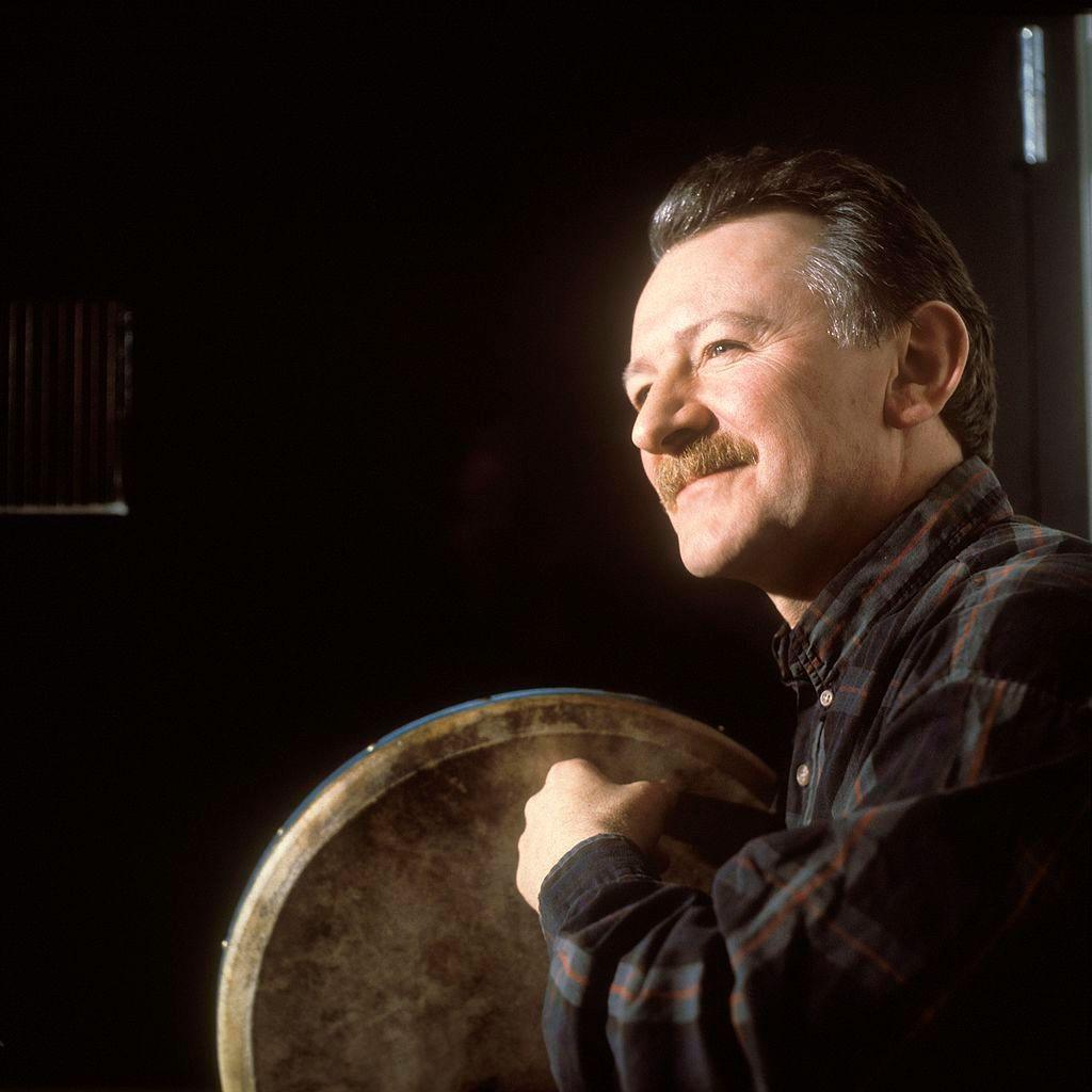 A close shot of a man in a grey shirt, with a moustache, playing the bodhran