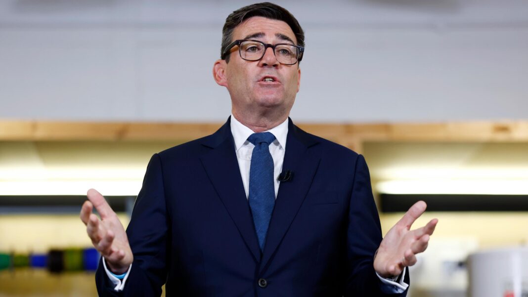 Andy Burnham wears a dark suit and blue tie and is mid speech, gesturing forwards with his hands
