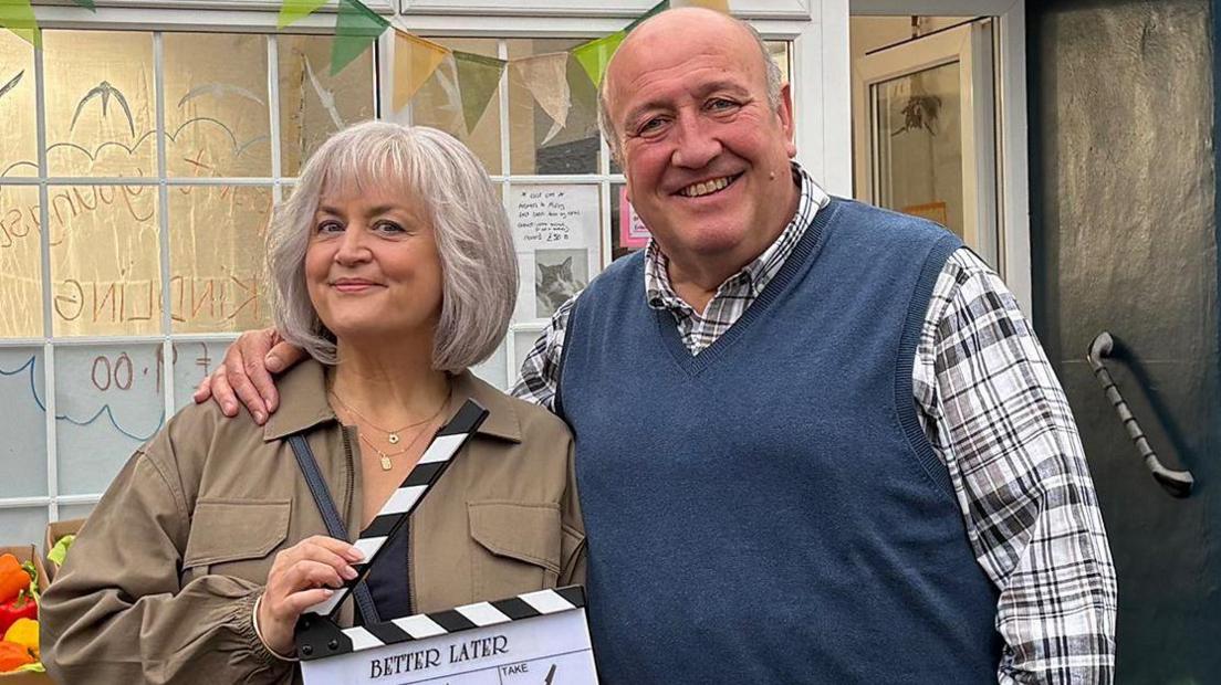 Ruth Jones stands with Steve Speirs with his arm around her. She is smiling holding a clapperboard, wearing a brown jacket has has a short grey bob. Steve Speirs is smiling, with a checked shirt with a blue jumper vest over it. A shop background is behind them, with a box of peppers in sight.