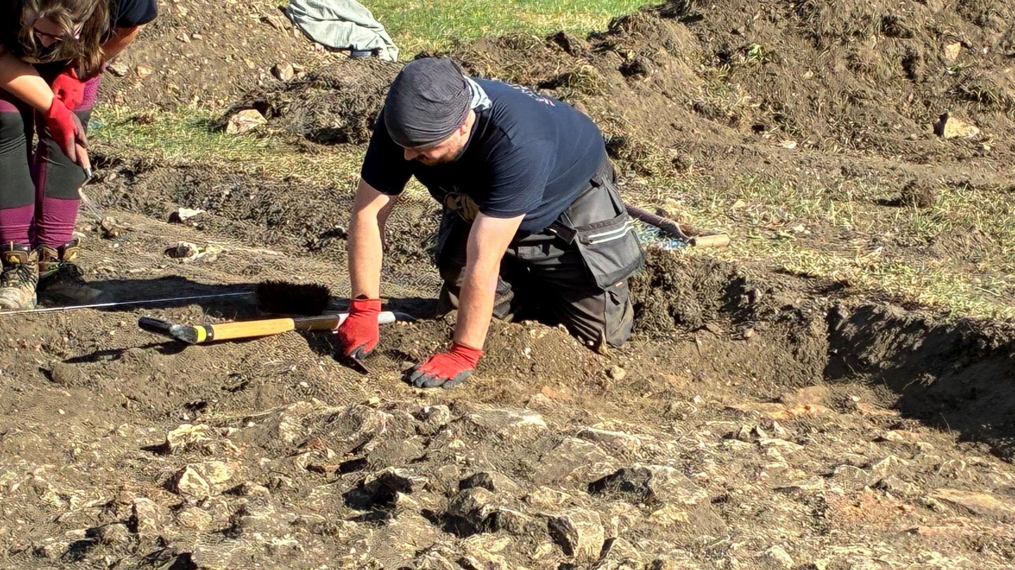 A man is knelt in the pit, scraping away at the soil in a red pair of gloves. The stones of the wall have been uncovered in front of him.