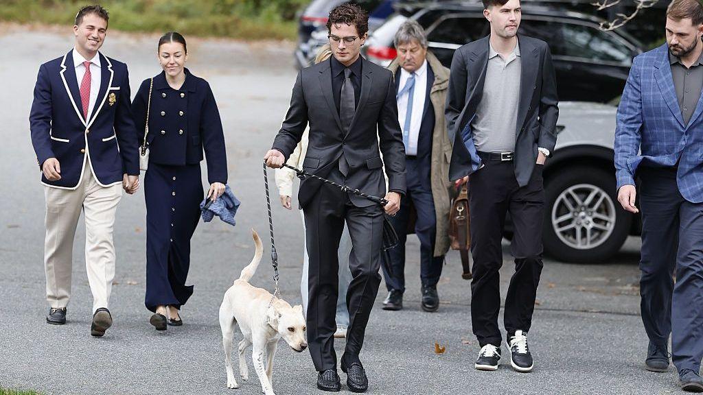 Braden Peters, known as Clavicular, is wearing a black suit and walking in a parking lot with entourage and a dog on a leash