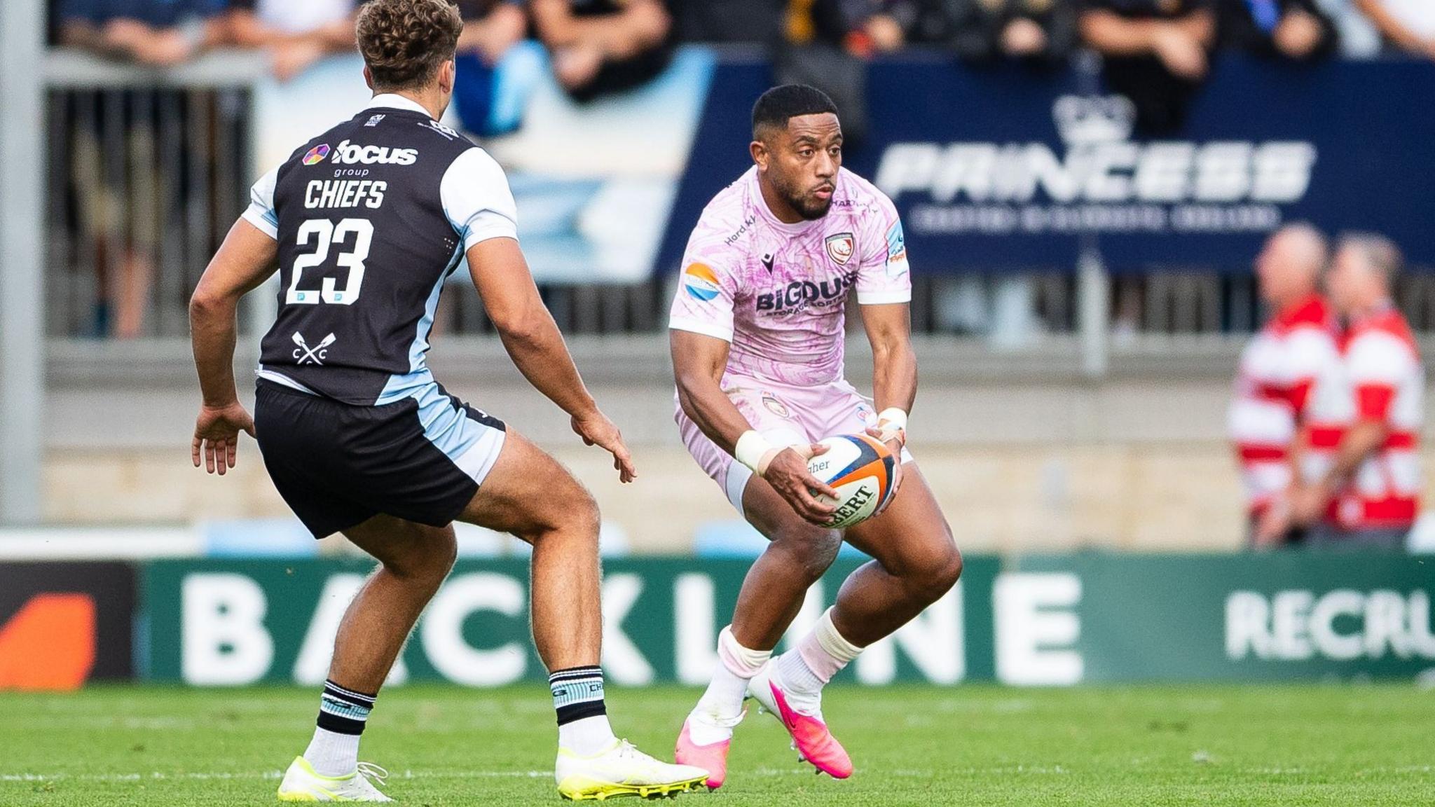 Phil Cokanasiga holds the ball in two hands low down as he moves on the pitch and Exeter's Zack Wimbush stands facing him