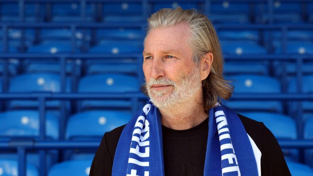 Robbie Savage in a black T-shirt poses with a blue Peterborough scarf