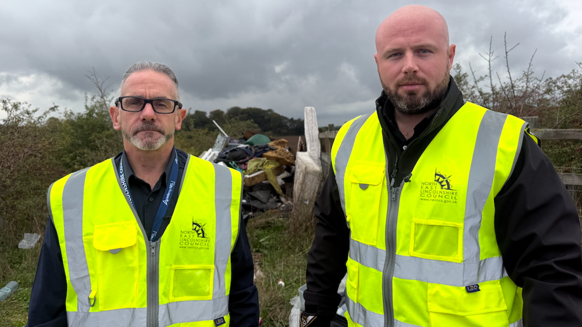 Anthony Horan and Ashley Hanley are both wearing yellow high vis jackets. They are stood front of a mound of fly-tipped waste including a mattress.