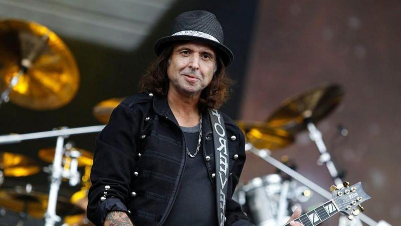 Phil, who has shoulder-length dark brown hair, and is wearing a black bucket hat, black t-shirt and black jacket. He is holding a guitar and is stood on a stage with a large drum set behind him.