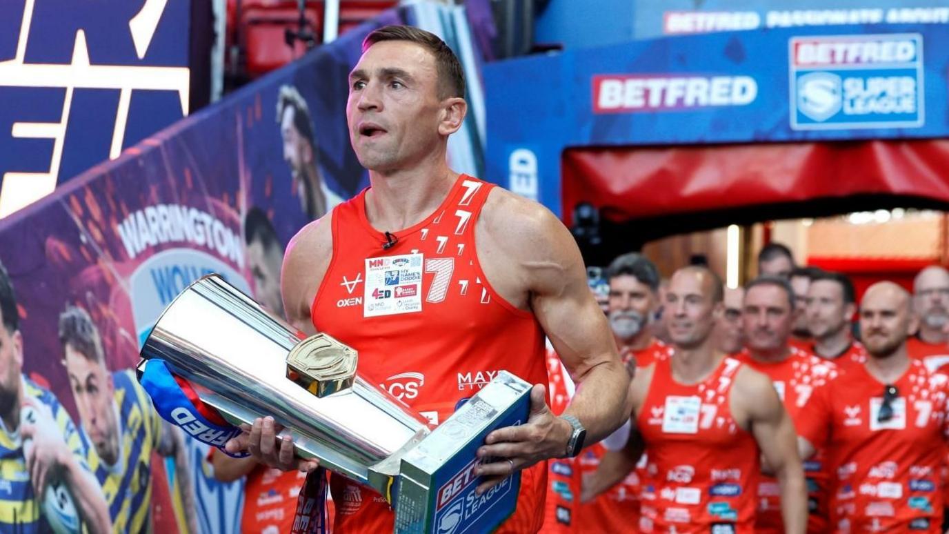 Sir Kevin Sinfield, wearing a red vest, carries a large silver trophy. Behind him is a team of men in red tops