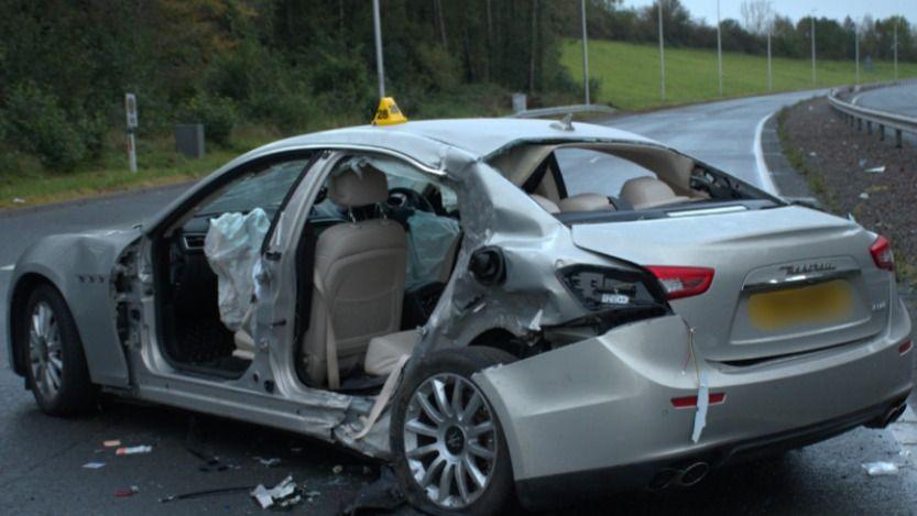 A behind view of a grey Maserati car in the middle of a lane on a dual carriageway. The car is visibly damaged with the airbags blown up and the doors missing. There is debris around the car on the road.