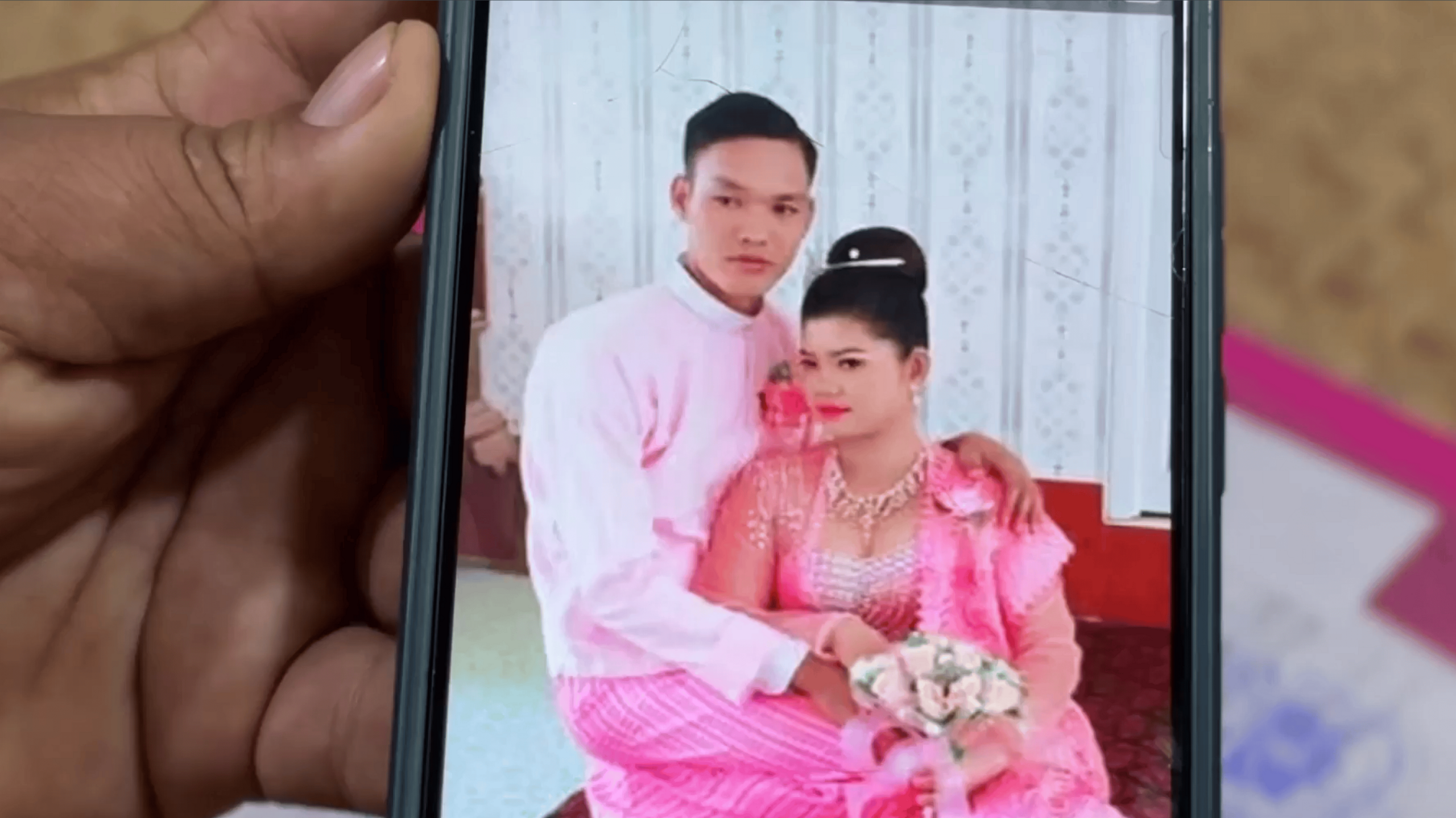 A hand holds a phone. On the screen are a man and a woman embracing in pink and white ceremonial clothing. The woman is holding a bouquet of white flowers.