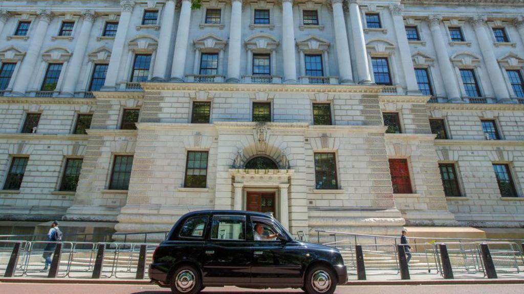 Taxi driving past the Treasury building in Whitehall - a pale brick Edwardian-style building. The first three floors and entrance steps can be seen here