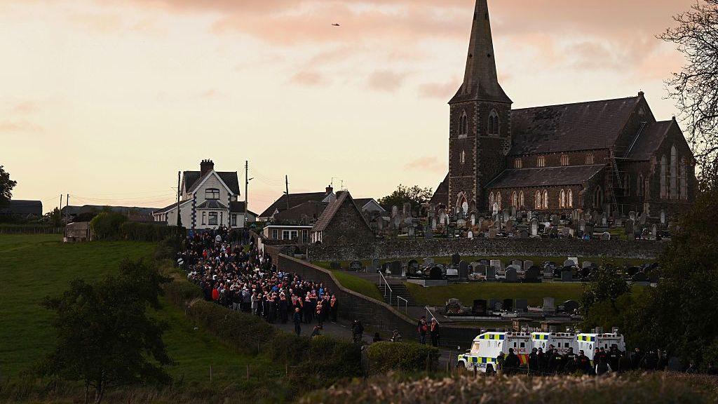 Orangemen gather on a road outside Drumcree Church, while police Land Rovers block the road ahead of them.
