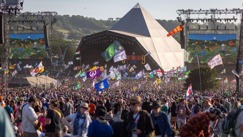 A large crowd in front of the Pyramid Stage at Glastonbury Festival. Lots of people are holding up large flags and waving them as the sun sets over the stage.