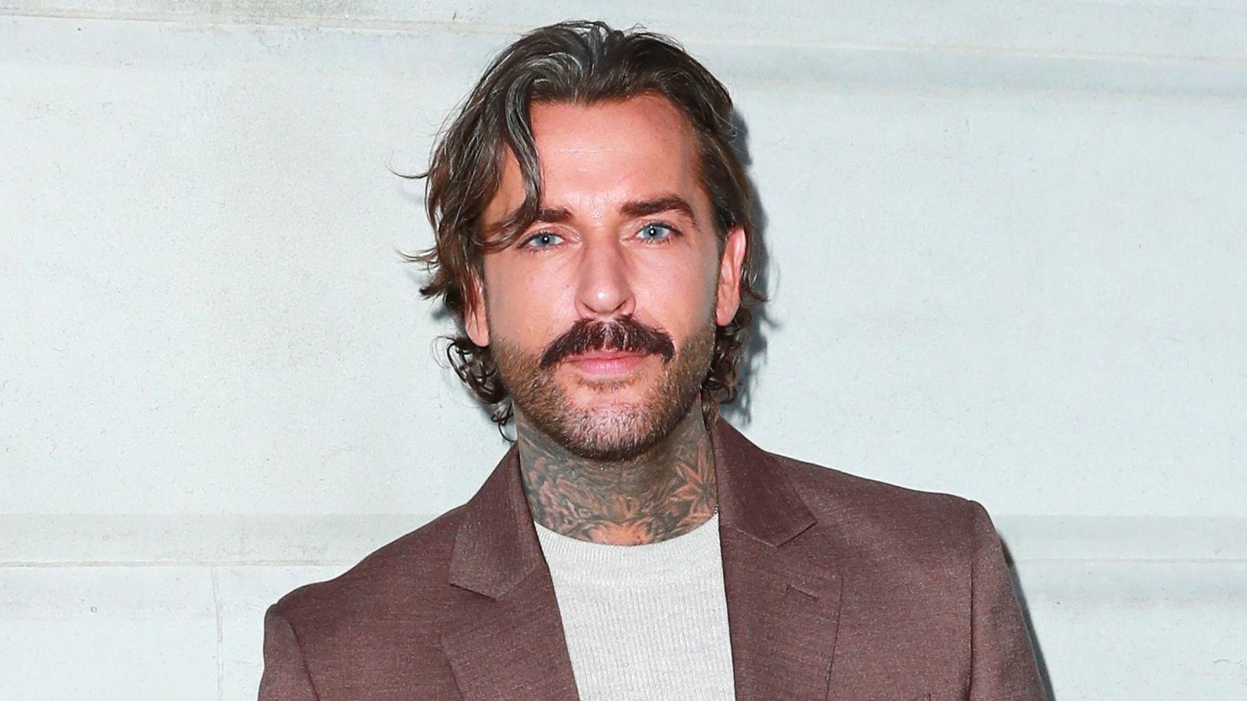Pete Wicks in a brown suit jacket with grey T-shirt looking at the camera
